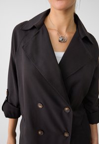 Chaqueta negra de doble botonadura con solapas en picos, mangas enrolladas y grandes botones marrones. Collar de plata con anillos entrelazados.