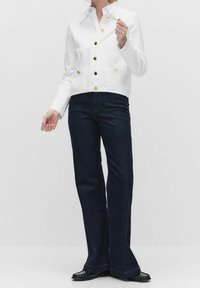 Bílá zkrácená bunda se zlatými knoflíky, která je kombinována s tmavě modrými zvonovými džíny. Hladká textura, dobře padnoucí střih a minimální akcenty.