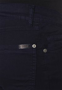 Jeans em denim azul-marinho com um bolso frontal com marca, fecho de botão metálico e detalhes de costura em contraste.