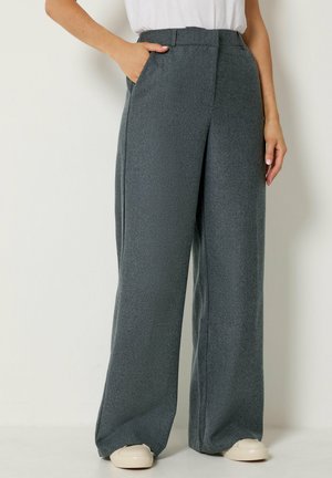Person trägt graue, hochgeschnittene, weit geschnittene Hose mit vorderen Falten, weiße Turnschuhe und ein in die Hose gestecktes weißes Hemd. Eine Hand in der Tasche.
