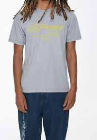 Lichtgrijs T-shirt van katoen met gele bedrukte tekst. Korte mouwen en een relaxed fit, gecombineerd met donkerblauwe spijkerbroek.