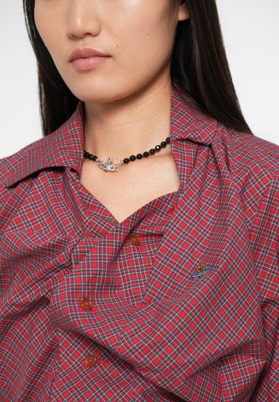 Camisa de botão xadrez vermelha com detalhes verdes e azuis, apresentando botões prateados e um pequeno logótipo bordado no tecido. Colar de contas pretas.