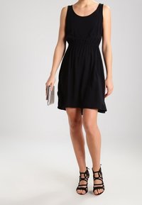 Schwarzes ärmelloses Kleid mit tailliertem Schnitt und ausgestellt geschnittenem Saum, kombiniert mit schwarzen Riemchensandalen und einer silbernen Clutch. Glatte Stoffoberfläche.
