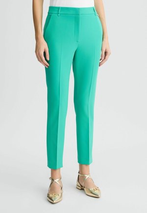 Woman wearing teal slim-fit ankle trousers paired with metallic gold pointed-toe flats with ankle straps.