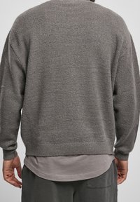 Mann in einem lockeren, strukturierten grauen Pullover über einem langen grauen Hemd, kombiniert mit dunklen grauen Hosen, von hinten gezeigt.