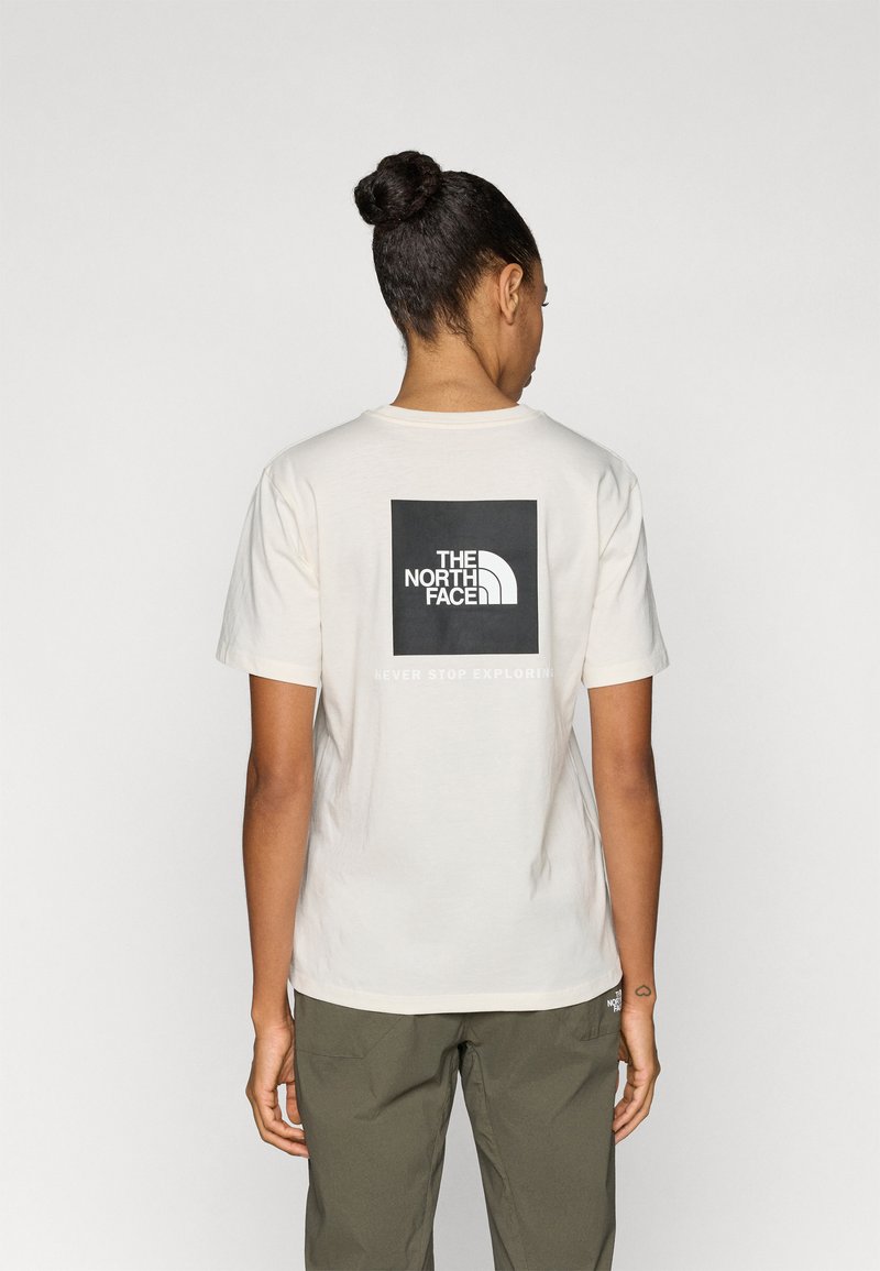 Βαμβακερό μπλουζάκι σε εκρού χρώμα με κοντά μανίκια, που φέρει ένα έντονο μαύρο τετράγωνο λογότυπο στην πλάτη και το κείμενο «NEVER STOP EXPLORING» από κάτω.