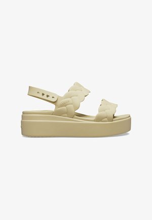 Sandales Crocs beiges avec un design de bride tressée, une bride arrière ajustable et une semelle épaisse pour plus de hauteur.