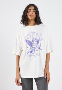 Balta per didelė t-shirt su purpuriniu grafiniu vaizdu cherubino su sparnais, tekstu "Ne tavo ANGELAS," ir "Širdies lūžių klubas" po juo.