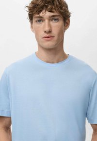 Mladý muž se světlými hnědými kudrnatými vlasy a modrýma očima, oblečený v světle modrém tričku s kulatým výstřihem, na jednoduchém bílém pozadí.