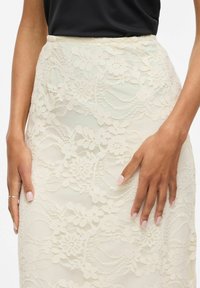 Mains reposant sur une jupe en dentelle florale couleur crème portée avec un haut noir, gros plan sur la taille et les hanches, montrant le motif de dentelle complexe.