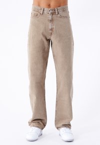 Pantalones vaqueros de denim color beige claro con corte recto, cierre de botón y cremallera, cinco bolsillos y una textura sutilmente desgastada. Zapatillas blancas mostradas abajo.