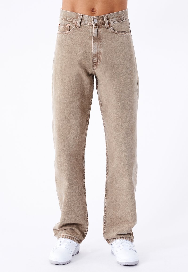 Pantalones vaqueros de denim color beige claro con corte recto, cierre de botón y cremallera, cinco bolsillos y una textura sutilmente desgastada. Zapatillas blancas mostradas abajo.