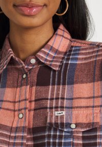 Camicia di flanella a quadri nei toni dell'arancione, marrone e blu. Presenta un colletto button-down e una tasca sul petto con etichetta. Texture morbida.