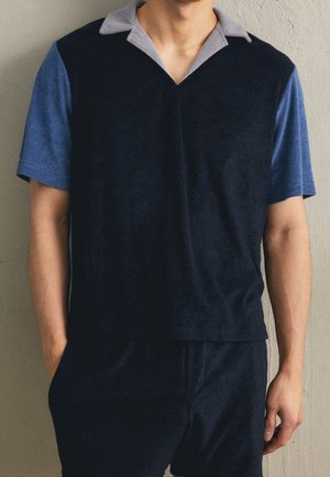 Homme portant une chemise noire à manches courtes avec des manches bleues et un col gris, debout devant un mur beige uni.
