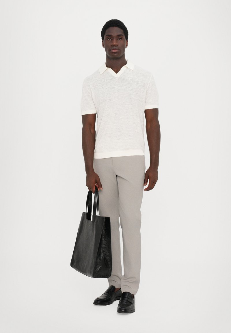 Polo shirt blanc à manches courtes avec un col en V, pantalon ajusté gris clair et un sac fourre-tout en cuir noir mat avec deux poignées. Mocassins noirs.