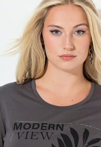 Vaaleatukkainen nainen sinisin silmin, jolla on ketjupainikkeiset korvakorut ja harmaa paita, jossa lukee "MODERN VIEW" ja jossa on lehtikuvio.