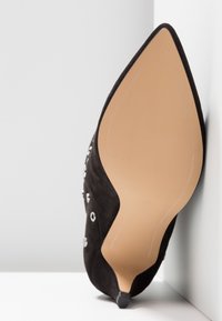 Chaussure à talons hauts en suède noir avec un bout pointu, dotée d'accents métalliques et d'un talon élancé. Semelle de couleur crème avec un détail de logo en relief.