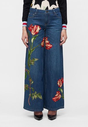 Blå wide-leg denim jeans med rød og hvid blomsterbroderi på forbenene, båret med sorte hæle.