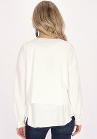 Weiße Bluse mit langen Ärmeln und geschichteter Gestaltung, mit abgerundetem Saum und geknöpften Manschetten, kombiniert mit dunklen Jeans. Angenehme Struktur.