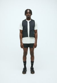Vesta fără mâneci de culoare neagră, din material ușor, cu fermoar frontal, purtată cu un tricou gri deschis, pantaloni scurți negri și adidași negri cu șosete.