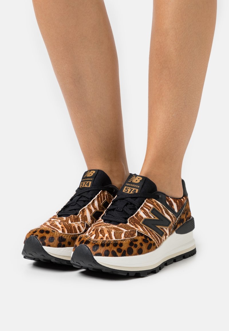 Jambes portant des baskets New Balance 574 avec un imprimé animal audacieux en marron et noir, semelles intermédiaires blanches et lacets noirs sur fond blanc.