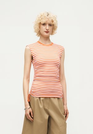 Άτομο με ξανθά σγουρά μαλλιά, ντυμένο με πορτοκαλί και λευκό ριγωτό αμάνικο τοπ και μπεζ παντελόνια με φαρδιά πόδια, στέκεται μπροστά σε απλό φόντο.