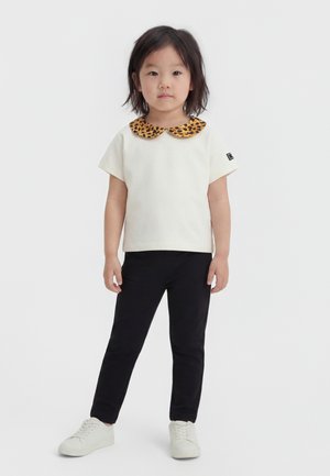 Bambino piccolo in piedi, indossa una maglietta bianca a maniche corte con colletto Peter Pan con stampa leopardata, pantaloni neri e scarpe da ginnastica bianche, su sfondo bianco.