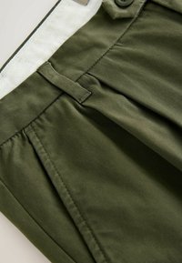 Olive green trousers made from smooth fabric feature front pleats, a button closure, and a contrasting waistband with a herringbone pattern.