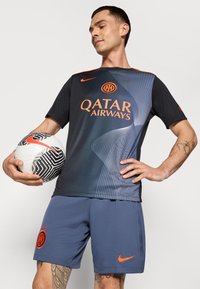 Svart och grå sporttröja med orange detaljer, med ett vågmönster och logotyp. Grå shorts med logotyp. Personen håller en mönstrad fotboll.