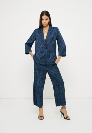 Kobieta stoi z rękami w kieszeniach, ubrana w granatową żakiet z kwiatowym wzorem o fakturze oraz pasujące szerokie spodnie, nosi czarne sandały na obcasie.