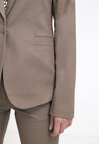 Hnědý blazer se hladkým, strukturovaným materiálem; má jednu kapsu vlevo a úzké rukávy, v kombinaci s odpovídajícími hnědými kalhotami.