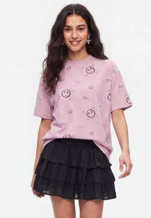 Kvinde med langt mørkt hår har en pink t-shirt med smilende ansigt og en sort flæset mini-nederdel på, smilende mens hun står foran en ensfarvet baggrund.