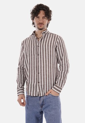 Uomo con capelli ricci che indossa una camicia marrone e bianca a righe verticali con bottoni e jeans blu, in piedi con una mano in tasca.