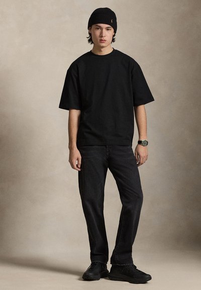 Νεαρός άνδρας που φοράει μαύρο σκουφί, μαύρο φαρδύ μπλουζάκι, μαύρο τζιν, μαύρα αθλητικά παπούτσια και ρολόι χειρός, μπροστά σε ένα απλό φόντο.