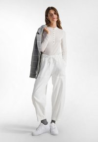 Maglietta bianca a maniche lunghe tipo henley con bottoni, abbinata a pantaloni bianchi larghi e sneaker bianchi. Giacca a scacchi grigia appoggiata sulla spalla.