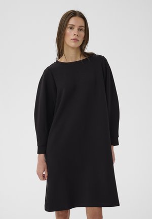 Junge Frau mit glatten braunen Haaren, die ein locker sitzendes, langärmliges, knielanges schwarzes Kleid vor einem schlichten hellen Hintergrund trägt.