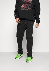 Svart sweatshirt med grafiskt tryck; ihop med svarta jeans med en logotyp; gröna och svarta sneakers med vita sulor.