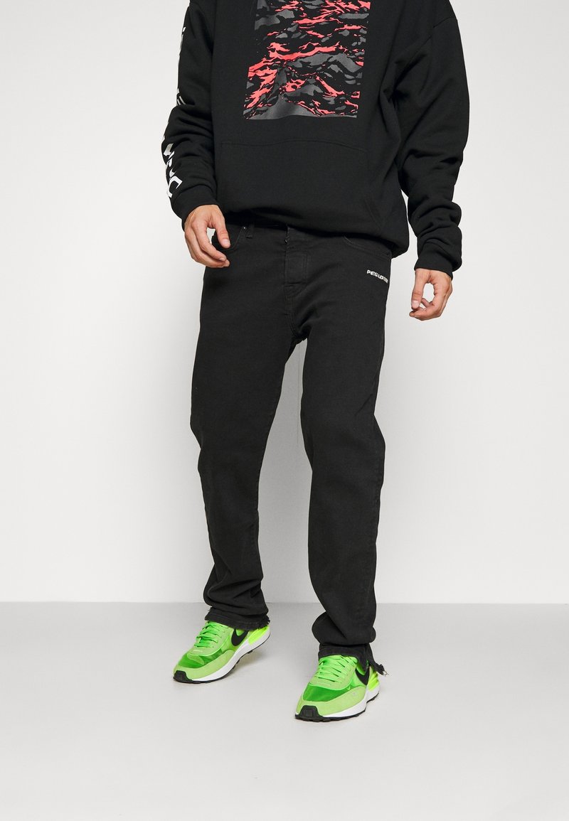 Svart sweatshirt med grafiskt tryck; ihop med svarta jeans med en logotyp; gröna och svarta sneakers med vita sulor.