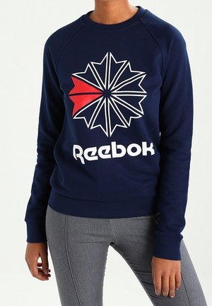 Osoba nosząca granatową bluzę Reebok z białym i czerwonym stylizowanym logo w kształcie gwiazdy oraz szare dopasowane spodnie.