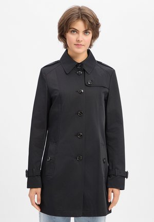 Femme aux cheveux courts portant un trench-coat noir boutonné avec poches, sangles à boucle sur les manches, debout devant un fond blanc uni.