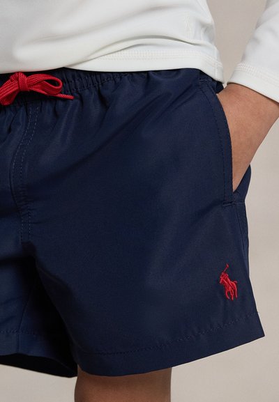 Kind trägt marineblaue Badeshorts mit rotem Tunnelzug und besticktem roten Polo-Spieler-Logo, Hand in der Tasche.