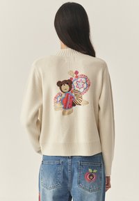 Krém színű kötött kardigán, hátul színes virágos részletekkel díszített hímzett medve mintával; bordázott szegély és mandzsetták. Farmernadrág almás folttal.