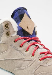 Beige suede sneaker with red speckled laces, navy checkered fabric at collar, and tan leather tag featuring "Reebok Classic" logo.