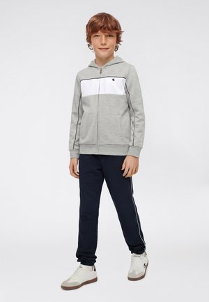 Junge mit rötlichem Haar, der einen grauen Kapuzenpullover mit Reißverschluss, marineblaue Jogginghosen mit weißen Streifen und weiße Turnschuhe trägt und vor einem schlichten Hintergrund steht.