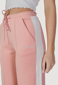 Pantaloni da tuta rosa chiaro con pannello laterale bianco, cintura elastica con cordino, tasca frontale e logo ricamato "GUESS 1981".