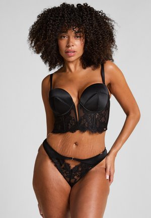 Vrouw met krullend haar in een zwart kanten lingerieset, poserend tegen een effen lichte achtergrond.