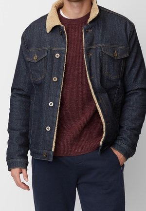 Mężczyzna w ciemnoniebieskiej kurtce dżinsowej z beżowym kołnierzem z polaru, pod którą ma bordowy sweter dziany i granatowe spodnie, z jedną ręką w kieszeni.