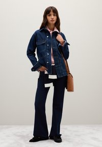 Giacca di jeans con due tasche sul petto, abbinata a una maglietta a righe e jeans a campana blu scuro. Borsa a tracolla marrone e scarpe nere.