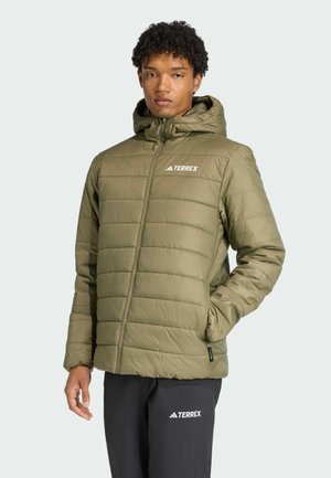Joven vestido con una chaqueta acolchada de color verde oliva con capucha y cierre delantero, de pie frente a un fondo claro.