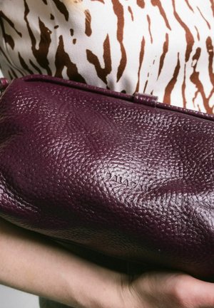 Clutch in pelle borgogna con superficie testurizzata, caratterizzato da un sottile logo impresso. Tenuto in mano contro una maglia a fantasie.
